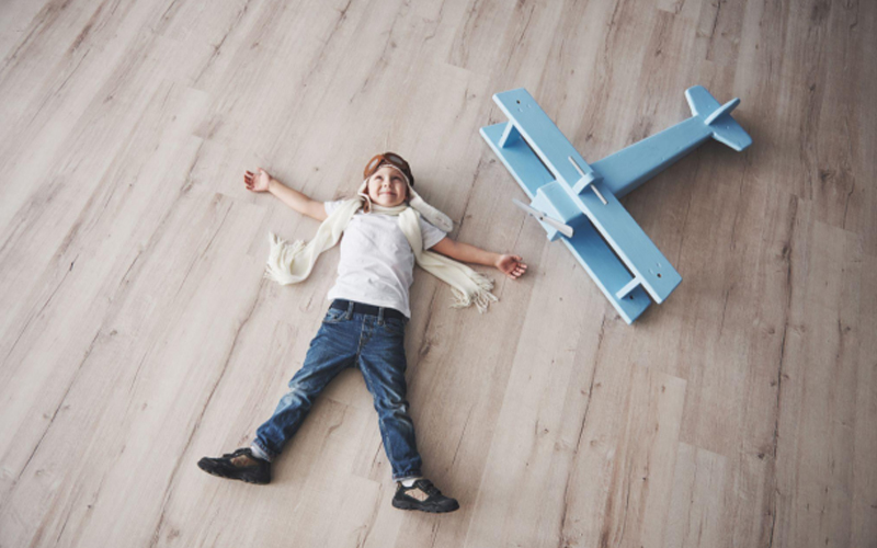 A little boy in pilot glasses lay on the vinyl wood look floor, beside a blue toy plane, dreaming of being a pilot when he grows up.